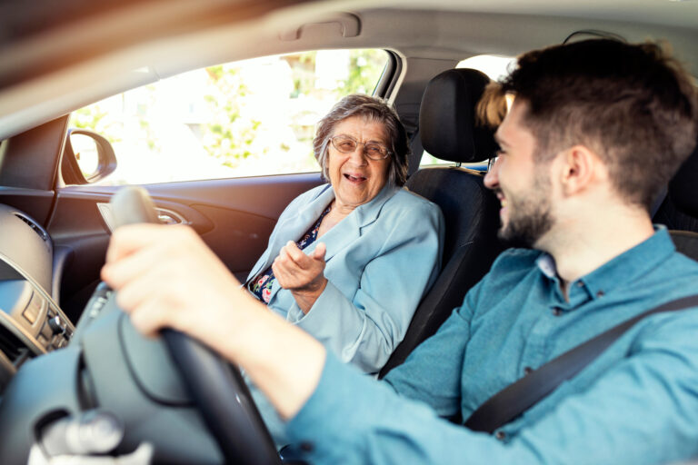 Ein junger Mann am Steuer eines Autos lacht gemeinsam mit einer älteren Dame, die auf dem Beifahrersitz sitzt.