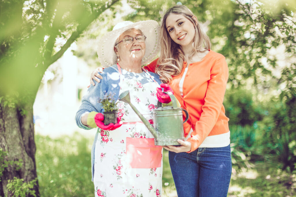 Seniorin und junge Frau stehen lächelnd mit Blumen und Gießkanne im Garten.